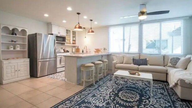 A modern kitchen with a central island, white cabinetry, and stainless steel appliances. There is a beige sofa and a patterned rug in the living area. - Cottage by the Sea steps to beach with heated pool (Carpinteria)