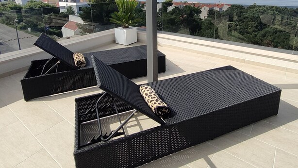 Terraza en la azotea con mobiliario de mimbre negro, incluyendo dos tumbonas y una pequeña mesa, con vistas a un paisaje urbano y al mar.