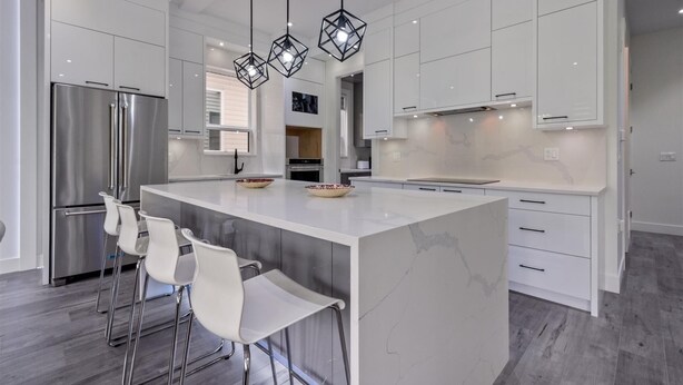 Une cuisine moderne dotée d’un îlot central, d’appareils électroménagers en acier inoxydable, de meubles blancs et de luminaires suspendus.