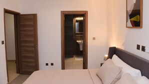 Un dormitorio con cama, mesita de noche, lámpara de noche, puerta y baño visible a través de una puerta abierta.