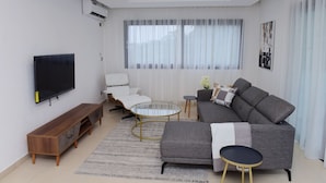 Un salón moderno con televisor de pantalla plana, un sofá gris, un sillón blanco, una mesa de centro de vidrio y un mueble de televisión de madera.