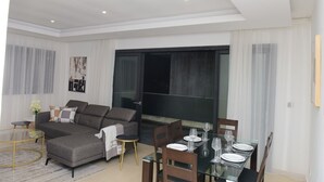 Una sala de estar moderna con un sofá seccional, una mesa de comedor de vidrio para cuatro personas y un gran ventanal corredizo que da a un área exterior.