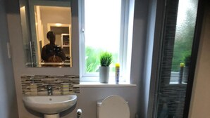 Ein Badezimmer im modernen Stil mit einem weißen Waschbecken, einer Toilette und einem Spiegel über dem Waschbecken. Ein Fenster bietet Blick auf Grünflächen, und der Boden hat ein schwarz-weißes Fischgrätmuster.