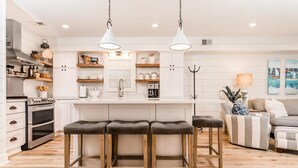 A modern kitchen with white cabinetry, a central island with stools, and a dining area with a sofa and a lamp.