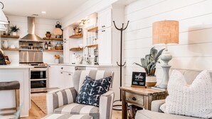 A modern kitchen with white cabinetry, stainless steel appliances, and wooden shelves. There is a striped armchair with a nautical pillow, a white sofa, and a small table with a plant and a sign.