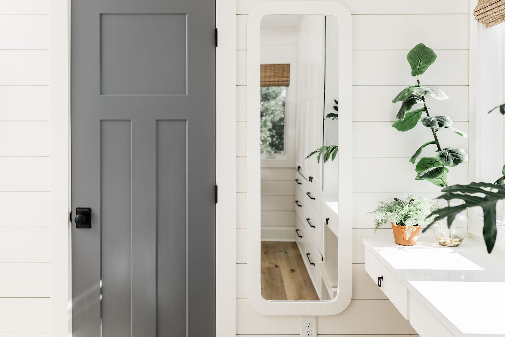 A modern bathroom with a grey door, a white vanity, and a mirror reflecting a hallway.