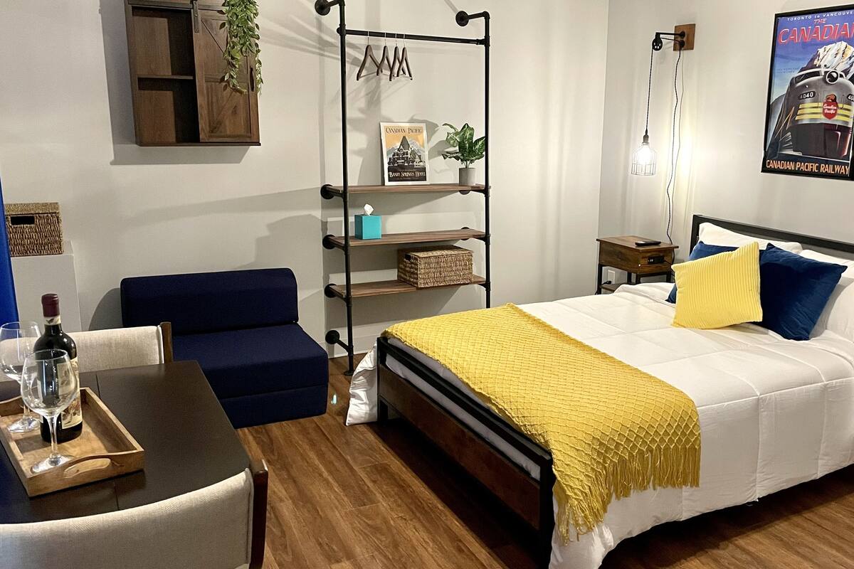 Ein Hotelzimmer mit einem Bett, einem Schreibtisch mit Stuhl, einem Regal mit Pflanze, einem Poster an der Wand und einem Fenster mit Vorhängen.