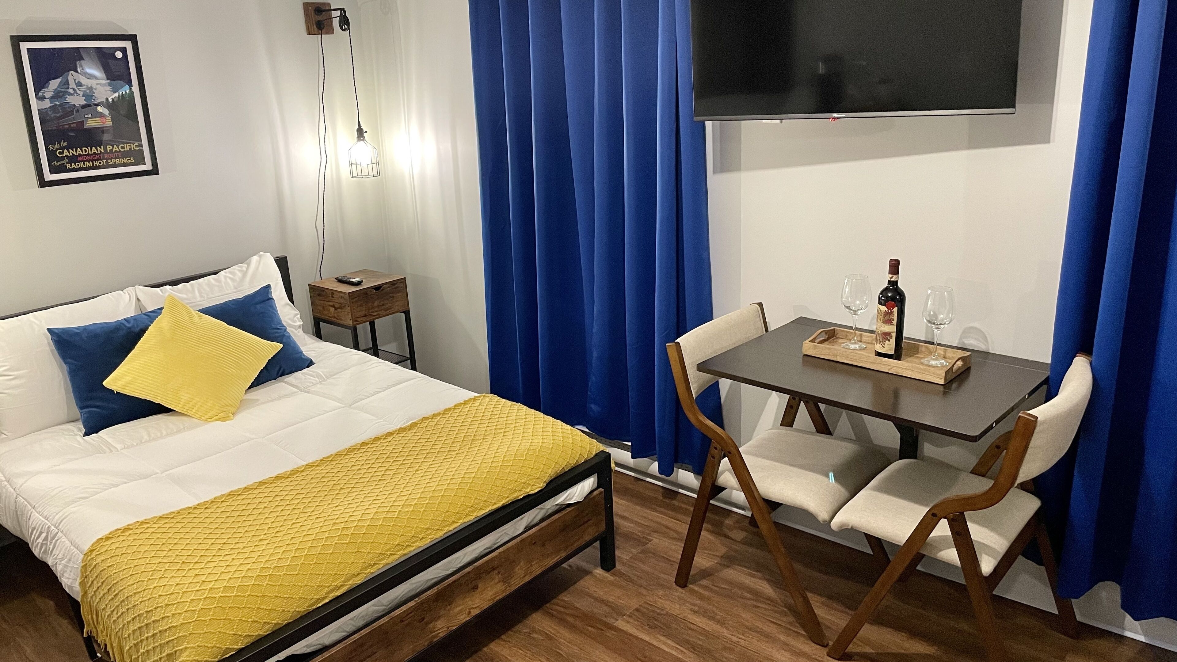 A hotel room with a bed, a small table with two chairs, a remote control on a nightstand, and a poster on the wall.