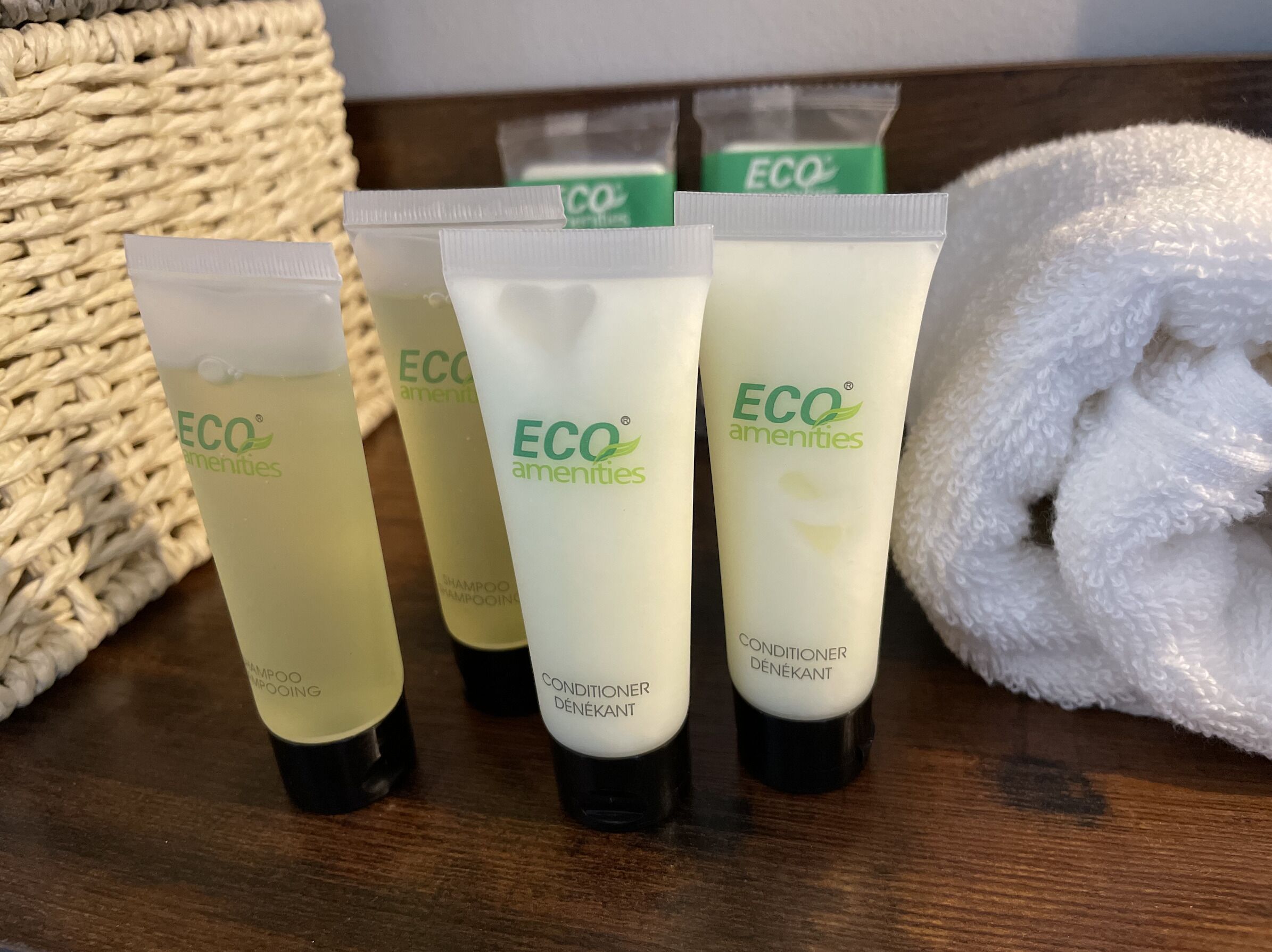Four bottles of ECO amenities shampoo and conditioner on a wooden surface, with a white towel and a woven basket in the background.
