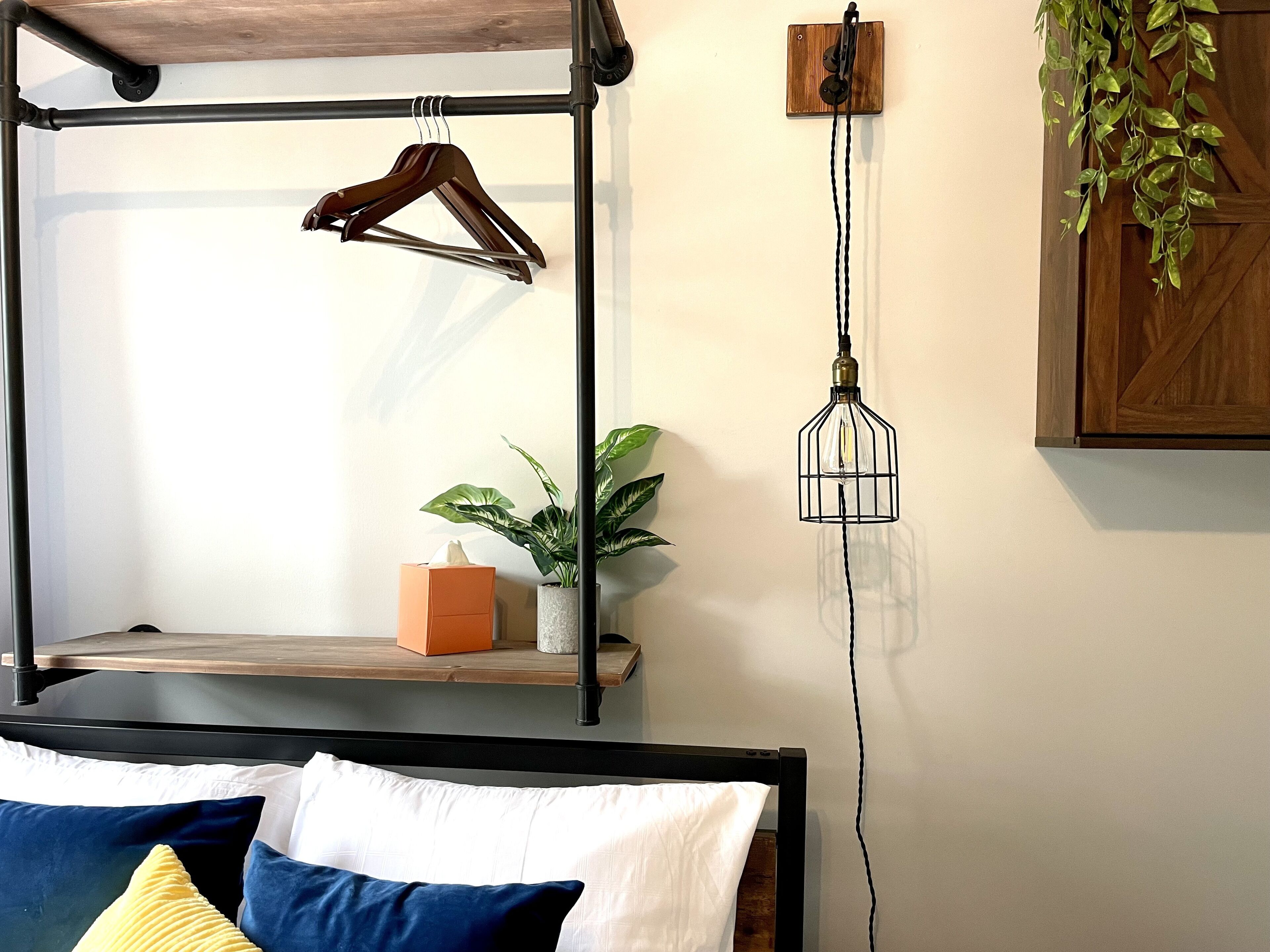 Uma cama com cabeceira de madeira, uma mesa de cabeceira com caixa de lenços, um candeeiro pendente e um vaso de planta.