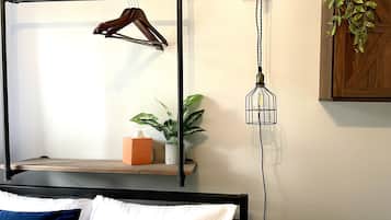 Uma cama com cabeceira de madeira, uma mesa de cabeceira com caixa de lenços, um candeeiro pendente e um vaso de planta.