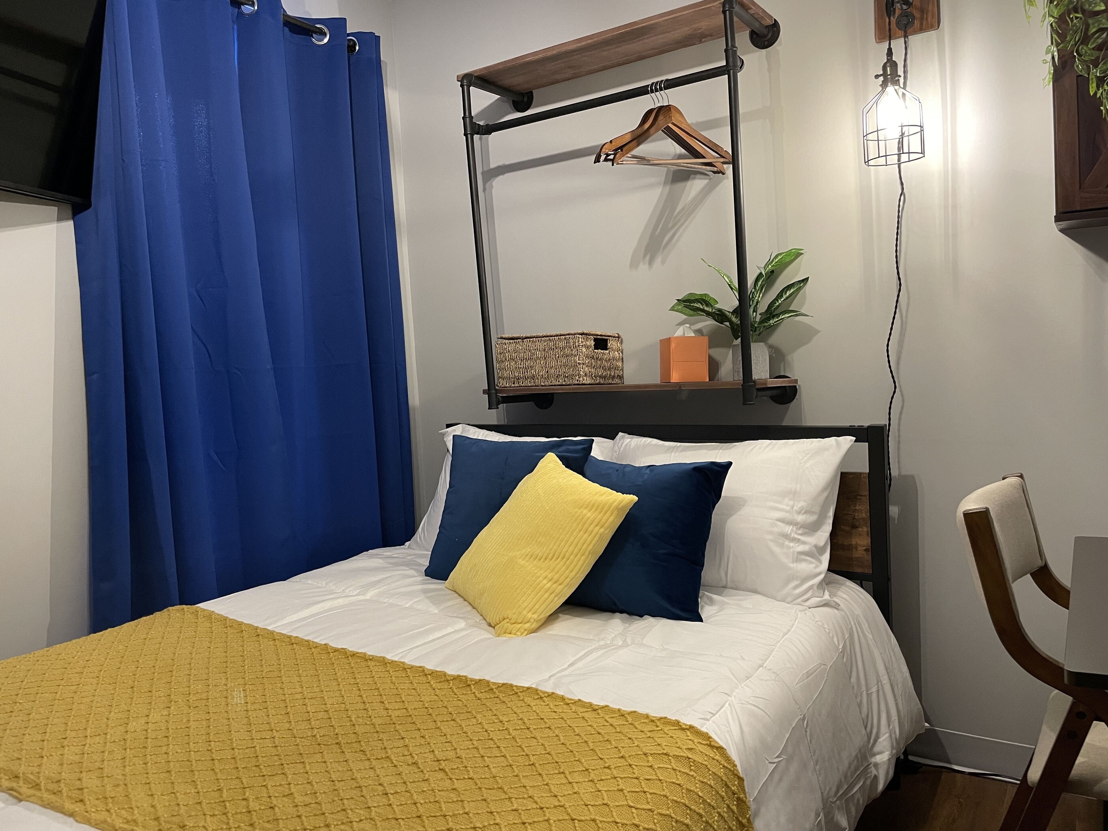 A bedroom with a bed, a wooden chair, a hanging light fixture, a shelf with a plant, and a blue curtain.