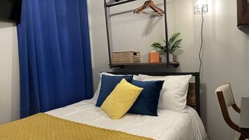 A bedroom with a bed, a wooden chair, a hanging light fixture, a shelf with a plant, and a blue curtain.