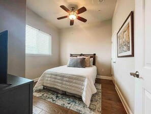 Ein Schlafzimmer mit Bett, Deckenventilator, Fenster mit Jalousien, Tür und einem gerahmten Bild an der Wand.