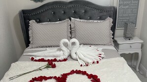 Un llit ben arrenjat amb roba blanca, dues tovalloles amb forma de cigne i un arranjament de pètals de rosa vermells en forma de cor sobre una manta blanca i esponjosa.