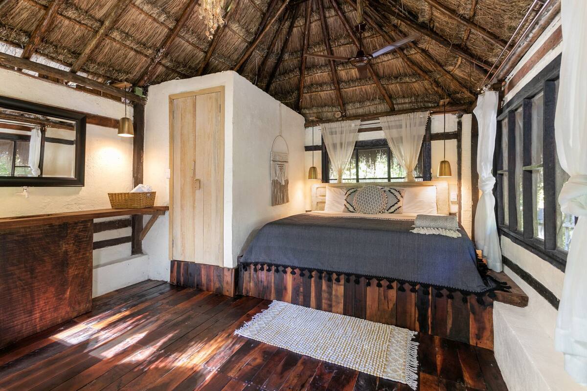 A rustic room with a wooden floor, a bed with a blue bedspread, a wooden wardrobe, a mirror, and a thatched roof.