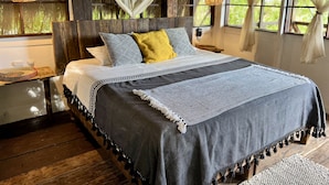 A bed with a grey and white patterned bedspread, a yellow pillow, and a grey blanket with tassels. There are two bedside tables with woven baskets and a power outlet. The room has wooden floors, a thatched ceiling, and windows with white curtains.
