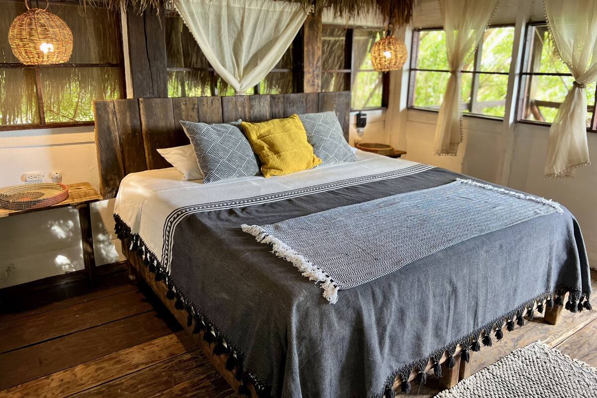 A bed with a grey and white patterned bedspread, a yellow pillow, and a grey blanket with tassels. There are two bedside tables with woven baskets and a power outlet. The room has wooden floors, a thatched ceiling, and windows with white curtains.