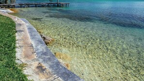 A clear view of a beach with a wooden pier extending into the water, a thatched-roof structure, and a grassy area.