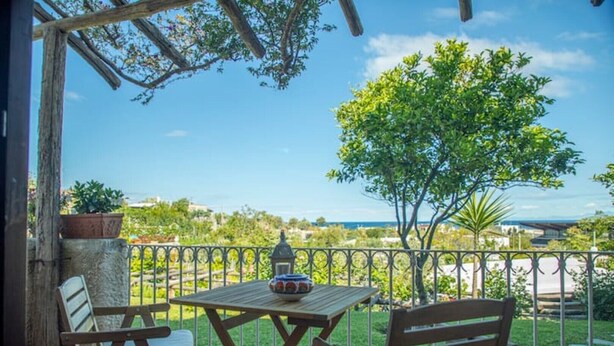 Un balcone con tavolo e due sedie, affacciato su un giardino rigoglioso e una vista lontana del mare.