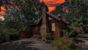 A log cabin with a chimney, surrounded by lush greenery and a paved driveway. - Hot Tub, Game Room, Pool Table, Fireplace | Minutes to Pigeon Forge (Sevierville)