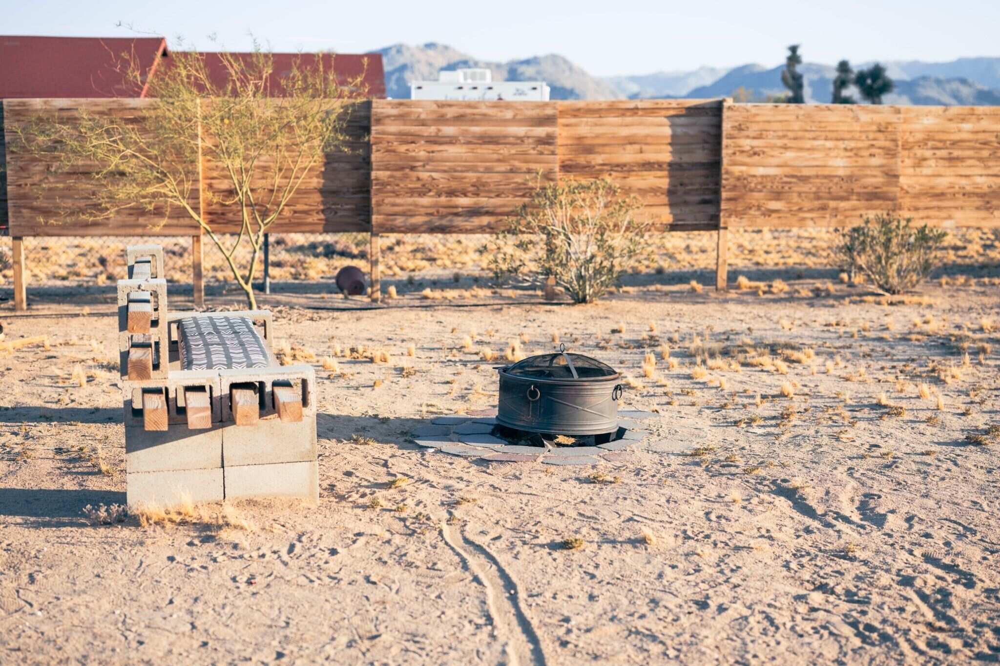 A desert campsite with a portable stove, a wooden bench, and a fence in the background.