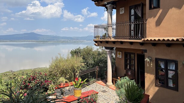 A two-story building with a balcony overlooking a lake, surrounded by potted plants and a stone-paved patio.