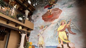 A chapel interior with a mural of angels, a wooden bench, and a glass-top table with a vase of flowers.