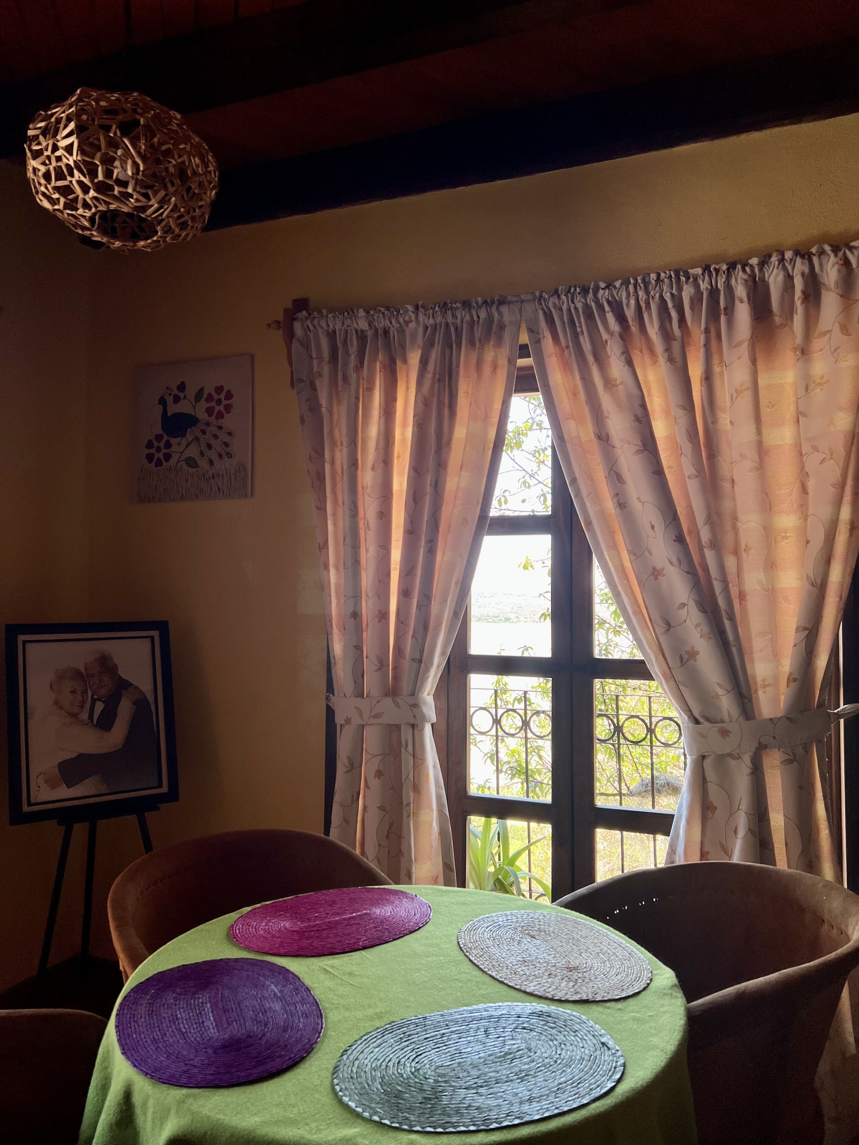 Une pièce avec une table recouverte d’une nappe verte, un tableau encadré au mur et une fenêtre avec des rideaux.