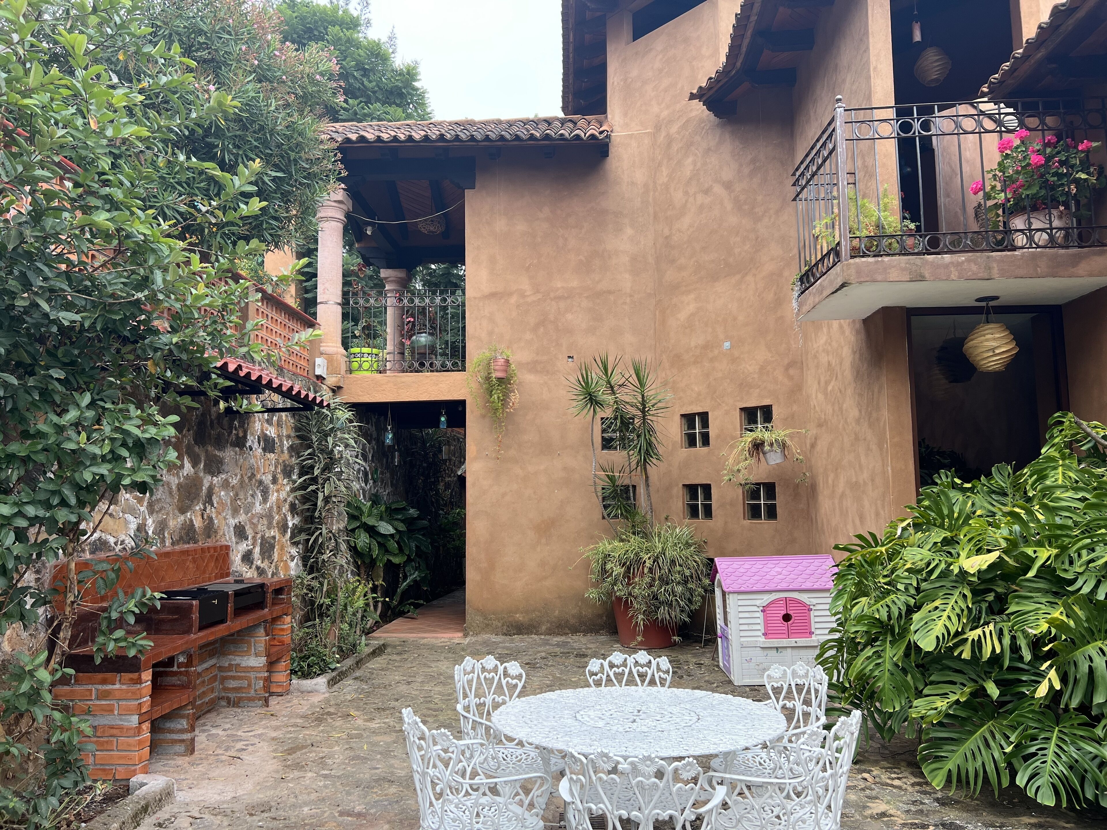 Une cour avec un barbecue en briques, une table et des chaises en métal blanc, des plantes en pot et une petite maison de jeu rose.