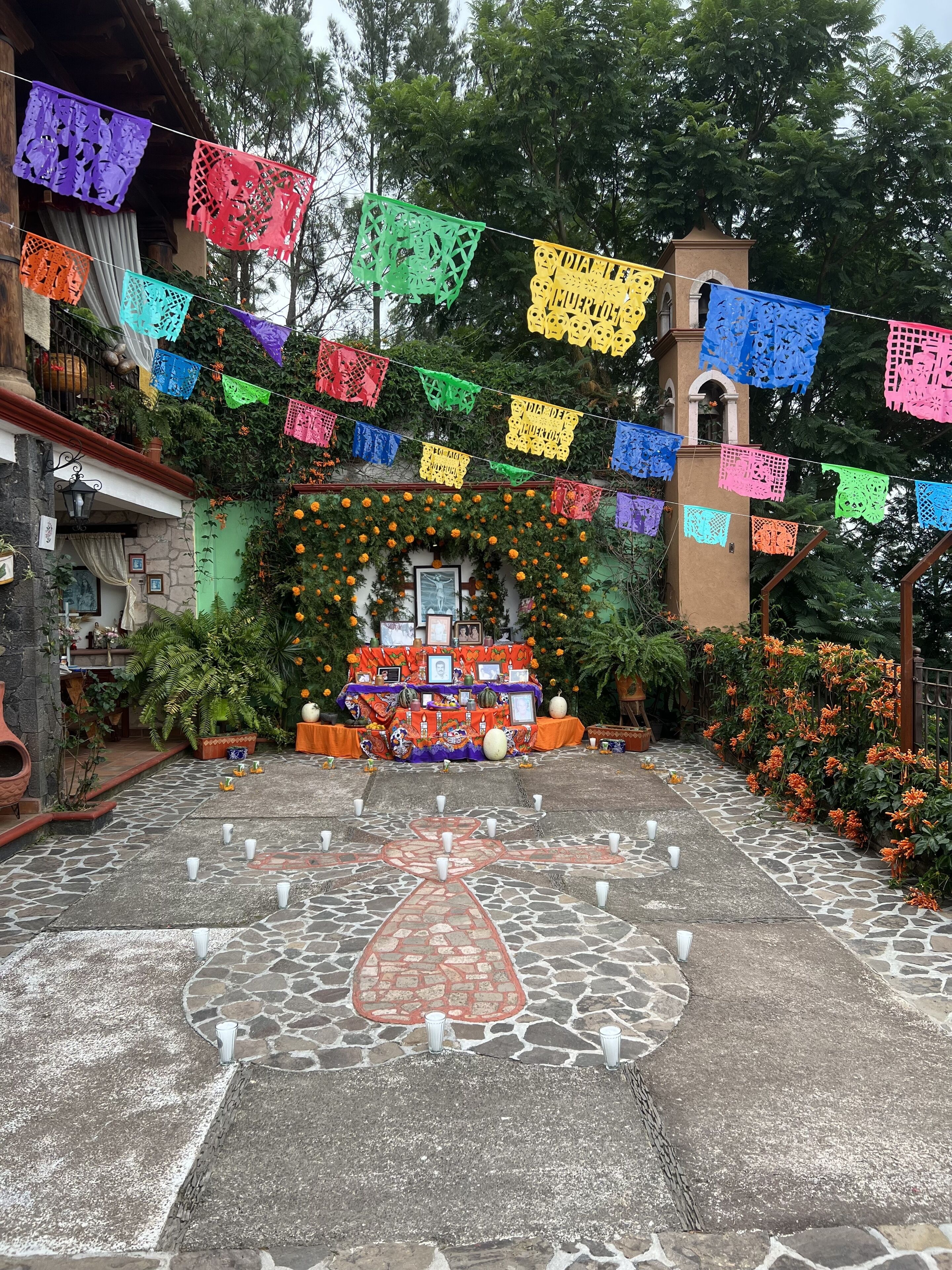 A courtyard with a decorated altar, surrounded by colorful papel picado banners, and a stone pathway leading to it.