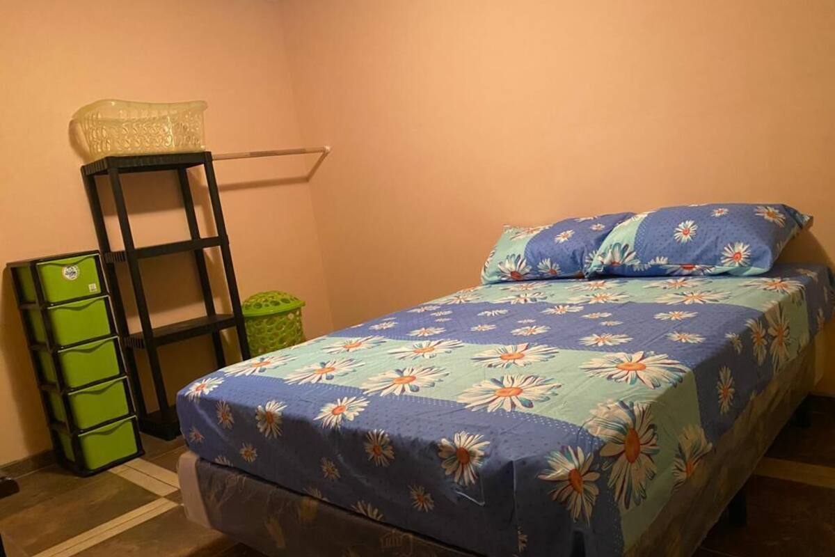 A bedroom with a bed covered in a floral bedspread, a green trash bin, a black shelf with a basket, and a green storage unit.