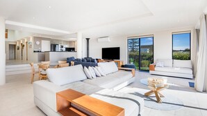 Una sala de estar moderna con un sofá, una mesa de centro y una televisión. Se ve una cocina al fondo y ventanas amplias que ofrecen vistas al exterior.