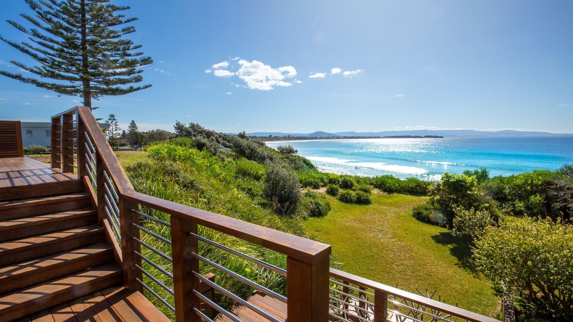 A wooden staircase leading to a deck overlooking a coastal landscape with a view of the sea and distant mountains.