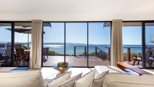 Una habitación moderna con grandes puertas de vidrio que dan a una terraza con vista al mar. Hay dos sofás y una pequeña mesa con una cesta.