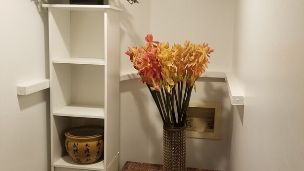 Dans un coin, on aperçoit une étagère blanche sur laquelle sont posés un pot de fleurs et un vase rempli de fleurs. En dessous de l’étagère, se trouve une commode en osier.