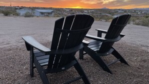 Dos sillas Adirondack mirando una puesta de sol sobre un paisaje desértico con edificios dispersos.