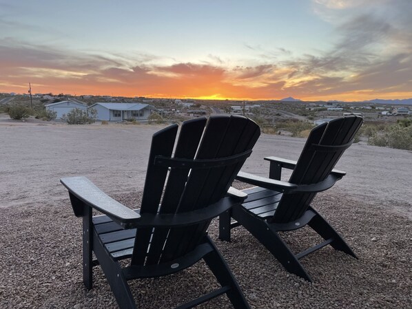 Zwei Adirondack-Stühle, die einem Sonnenuntergang über einer Wüstenlandschaft mit vereinzelten Gebäuden entgegenblicken.