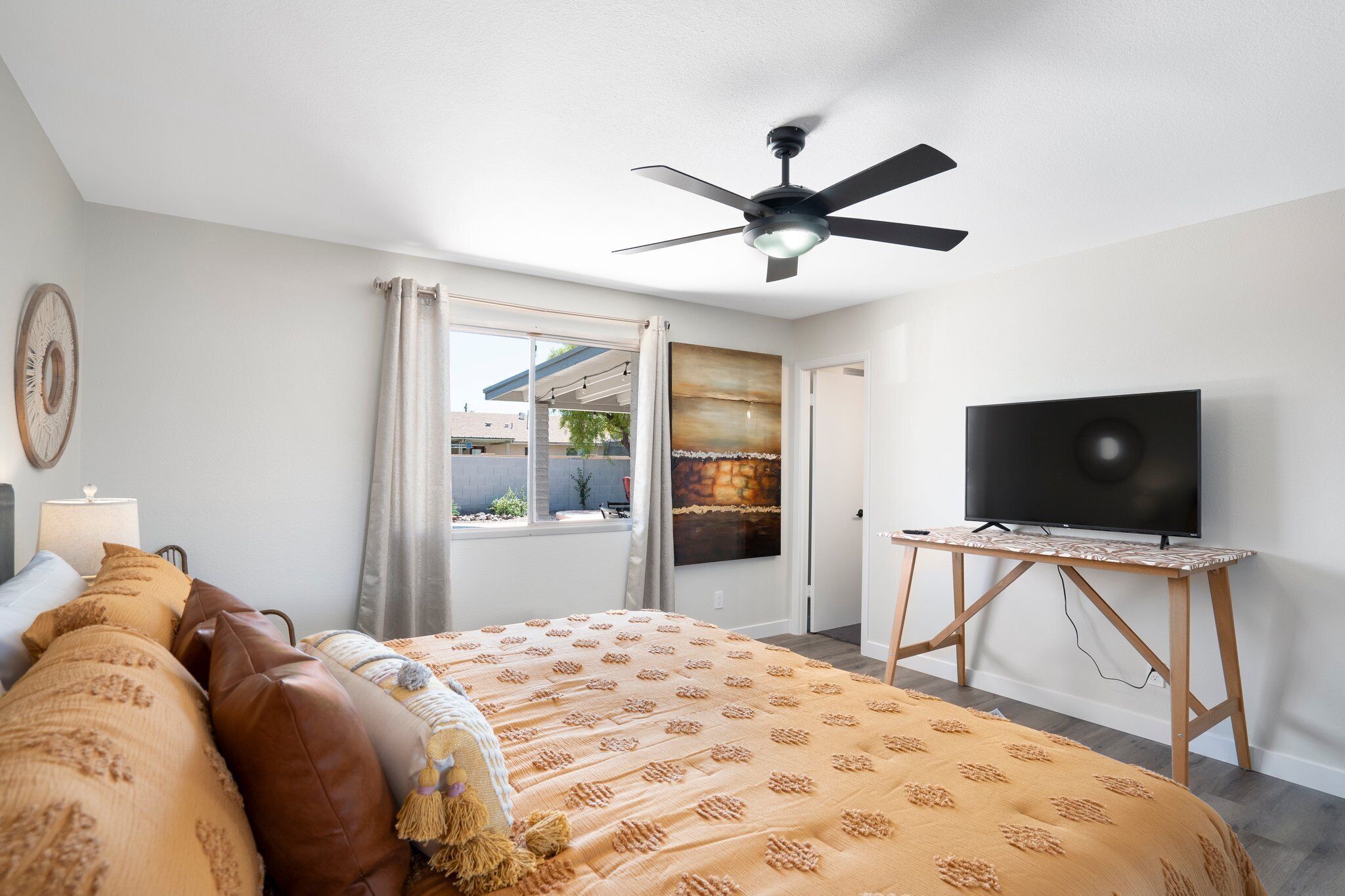 A bedroom with a bed, a ceiling fan, a TV on a wooden stand, and a window with curtains.