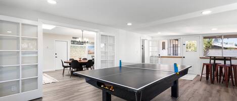 A modern indoor space with a ping pong table, a dining area, and a kitchen area.