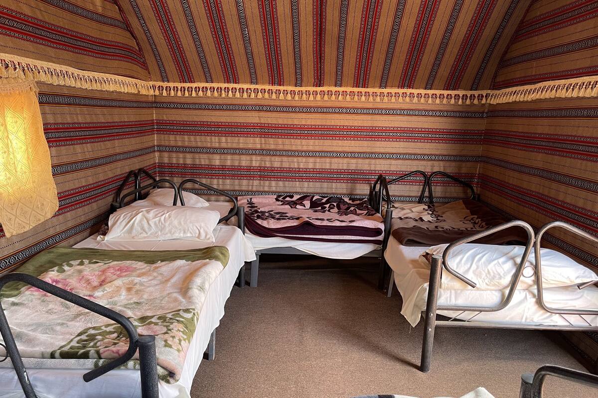 Une pièce avec quatre lits en métal, chacun recouvert d’une couvre-lit à motifs différents. Les murs de la pièce sont décorés de rayures et le sol est recouvert d’une moquette.