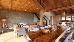A spacious living area with a wooden ceiling, a stone wall, a sofa set, a dining table with chairs, and a kitchen in the background. - Bain à Remous+Terrasse Panoramique! Chalet Raffiné (Les Deux Alpes)