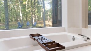 A modern bathroom with a bathtub, a window with a view of a forest, and a wooden tray on the tub. - Modern Poconos Game House! Hot Tub, Fire Pit (East Stroudsburg)
