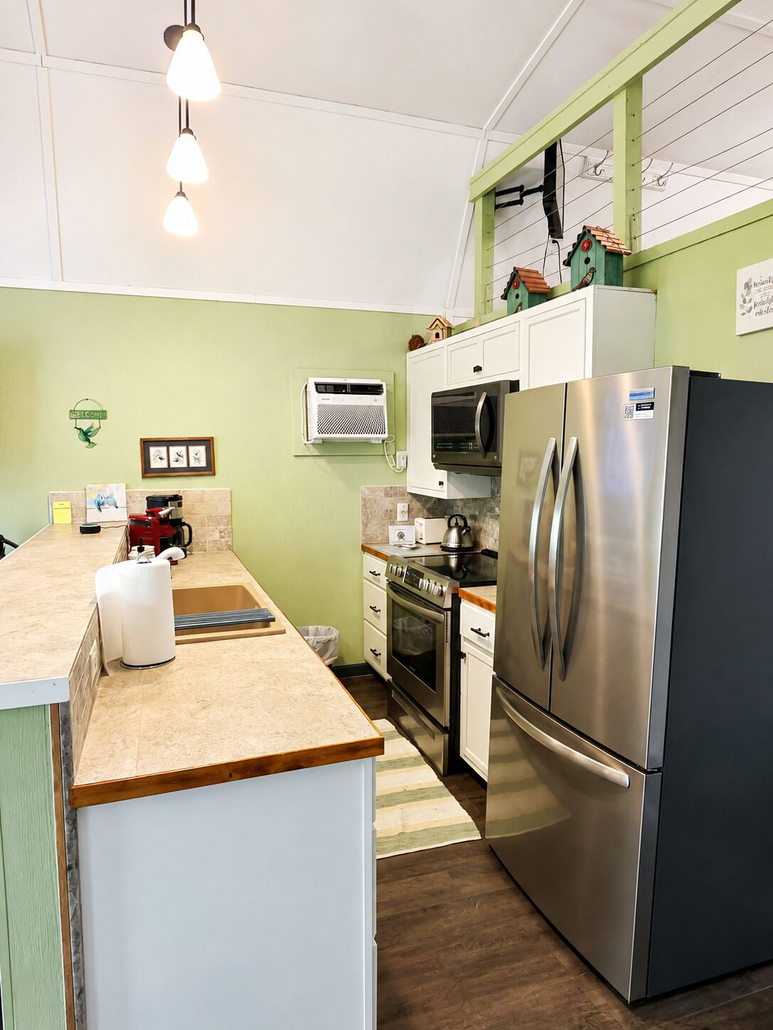 Cocina moderna con refrigerador de acero inoxidable, horno y microondas. Cuenta con encimera de madera y una pequeña isla con hervidor.