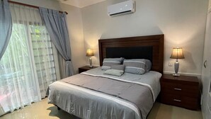 A bedroom with a bed, two bedside tables, a ceiling fan, an air conditioning unit, and a window with curtains. - Casa De Namaste- Relaxing 1 Br Condo near Playa's best Beaches & 5th Ave! (Playa del Carmen)