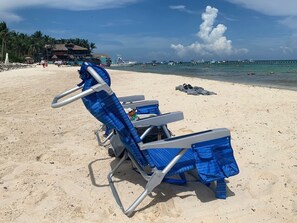 A beach with two blue sun loungers, a thatched-roof building, and a clear blue sky.
