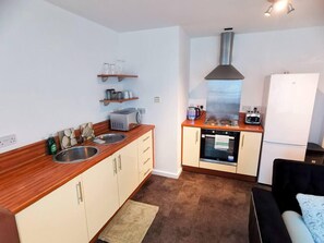 A compact kitchen with a sink, stove, oven, and a refrigerator. There are wooden shelves above the sink and a small countertop with a toaster and kettle.