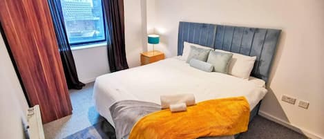 Hotellihuone, jossa on suuri sänky, yöpöytä lampun kanssa, ikkuna verhoilla ja näkymä kaupunkiin.