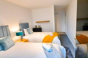 A hotel room with two beds, a nightstand with a lamp, a small table, and a wall-mounted shelf. - 2-bed Apartment Near City Centre (Salford)