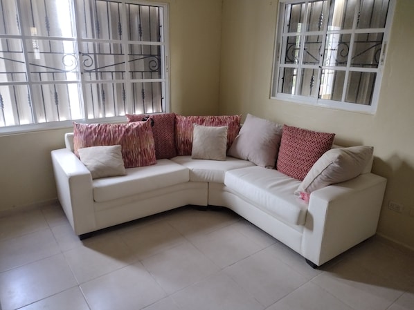 A white L-shaped sectional sofa with patterned cushions, positioned in a room with tiled flooring and two windows with metal bars.